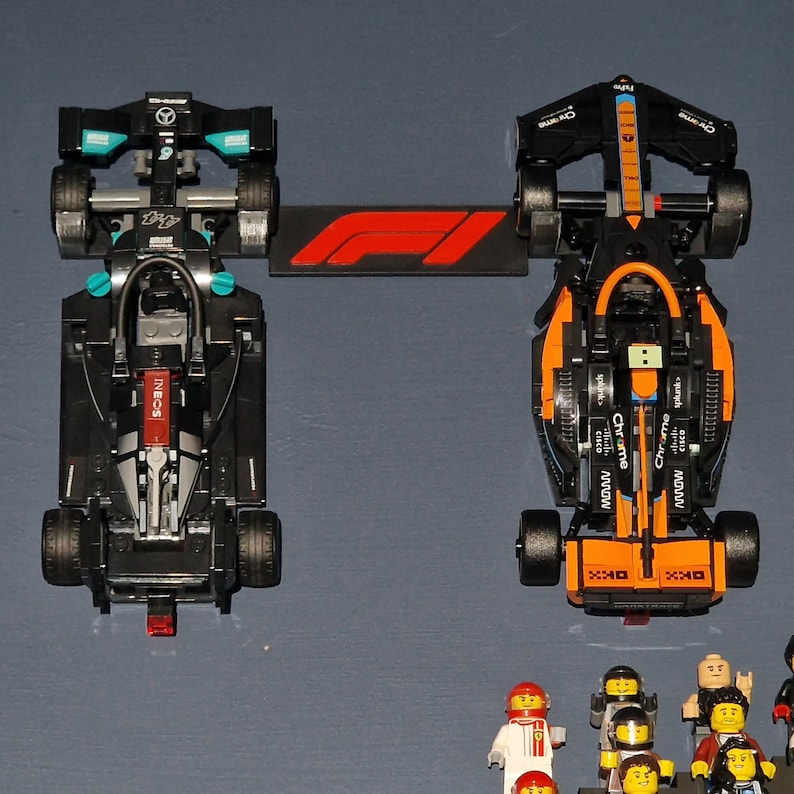 Puede incluir: Dos coches de carreras de F&oacute;rmula 1 negros con detalles en naranja y azul. Los coches est&aacute;n hechos de piezas de Lego y est&aacute;n sobre una superficie gris. Los coches se enfrentan y hay un logotipo de F&oacute;rmula 1 rojo y blanco entre ellos.