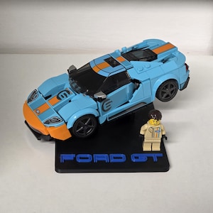 May include: A light blue and orange Ford GT model car with black wheels and a black base. The base has the words "FORD GT" in blue. A small figurine of a driver is standing next to the car.