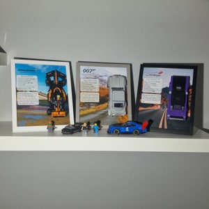以下が含まれることがあります： 情報テキスト付きの3つの額入りカーモデル。フレームにはマクラーレン、アストンマーティン、ダッジチャレンジャーが表示されています。小さなミニカーとフィギュアが白い棚のフレームの前に配置されています。