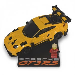 May include: A yellow toy sports car with black accents and a rear spoiler. The car sits on a black base with the text "GT3RS" in orange. A small figure in red clothing stands next to the car.