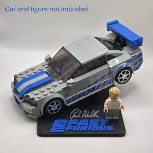 May include: A gray and blue building block model car with a rear spoiler, displayed on a black stand featuring the text 2 FAST 2 FURIOUS and a signature. A small plastic figure stands next to the car. The text Car and figure not included is visible.