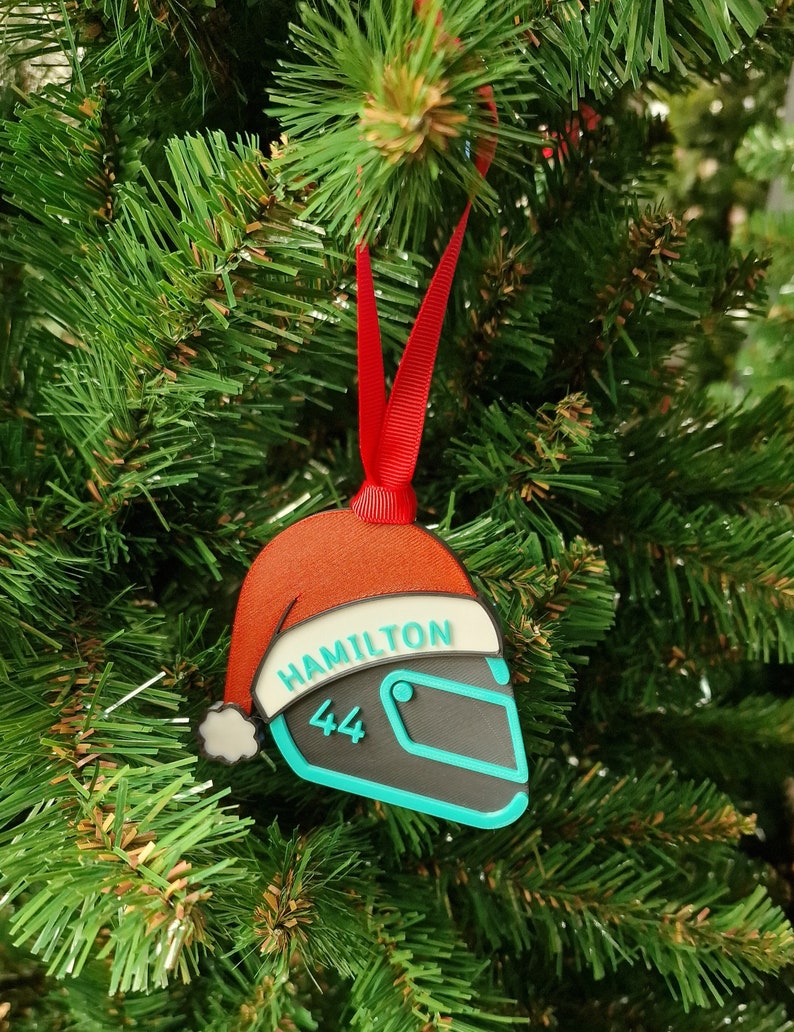Op de afbeelding: Een kerstboomversiering in de vorm van een racehelm, met een kerstmutsontwerp. De ornament is zwart, teal en rood, met de naam "HAMILTON" en het nummer "44". Het hangt aan een rood lint.
