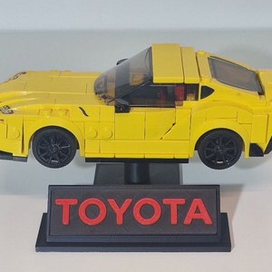 May include: A yellow Lego model of a Toyota Supra sports car on a black display stand with the word "TOYOTA" in red letters.