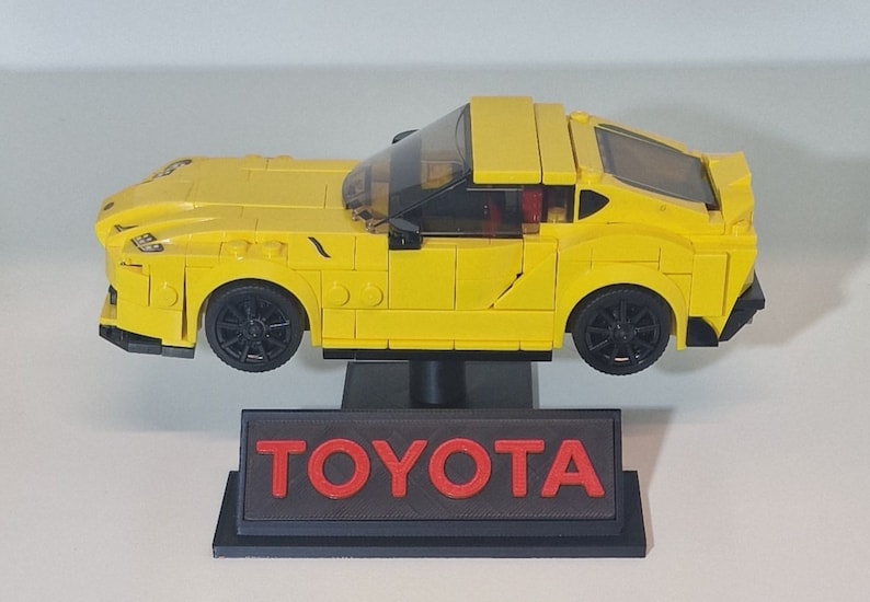 May include: A yellow Lego model of a Toyota Supra sports car on a black display stand with the word "TOYOTA" in red letters.
