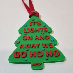 Puede incluir: Adorno navide&ntilde;o verde con forma de &aacute;rbol, con el texto rojo "IT'S LIGHTS ON AND AWAY WE GO HO HO HO". Tiene una cinta roja para colgar y est&aacute; decorado con peque&ntilde;os detalles azules y grises. Un logotipo de F&oacute;rmula 1 en la parte inferior.