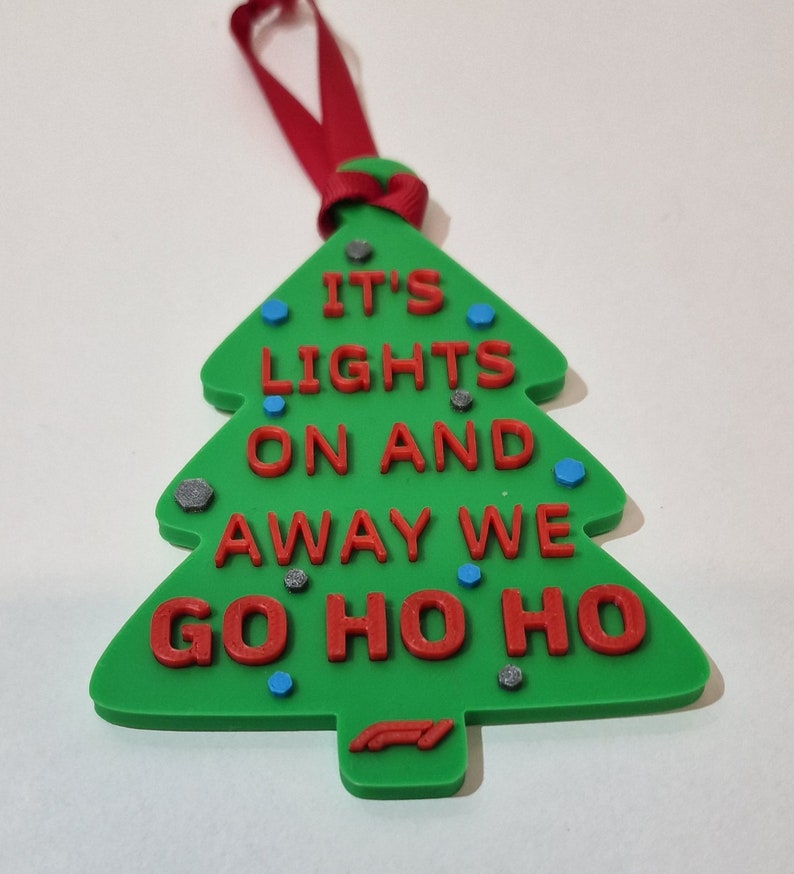 Puede incluir: Adorno navide&ntilde;o verde con forma de &aacute;rbol, con el texto rojo "IT'S LIGHTS ON AND AWAY WE GO HO HO HO". Tiene una cinta roja para colgar y est&aacute; decorado con peque&ntilde;os detalles azules y grises. Un logotipo de F&oacute;rmula 1 en la parte inferior.