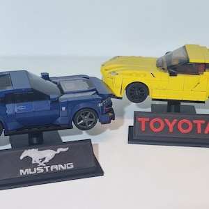 May include: Two toy car models on display stands. A blue Ford Mustang model with a black spoiler and a yellow Toyota Supra model. The stands have the car manufacturer's logos printed on them.
