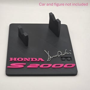 May include: A black rectangular display stand featuring two upright supports, pink raised lettering reading HONDA S2000, and a grey signature. The text Car and figure not included is printed in pink at the top of the image.