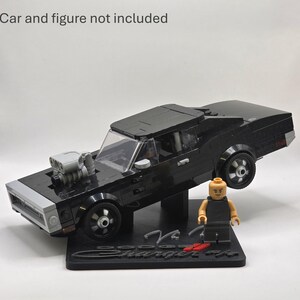 May include: A black building block model of a Dodge Charger car with a grey engine protruding from the hood, mounted on a black display stand with the text Dodge Charger R/T. A small plastic figure stands on the base. Car and figure not included.