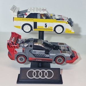 May include: Two Lego model race cars, one yellow and white with the number 5, and one gray and red, are displayed on a black stand. The stand features the Audi logo. The cars are detailed with racing decals.