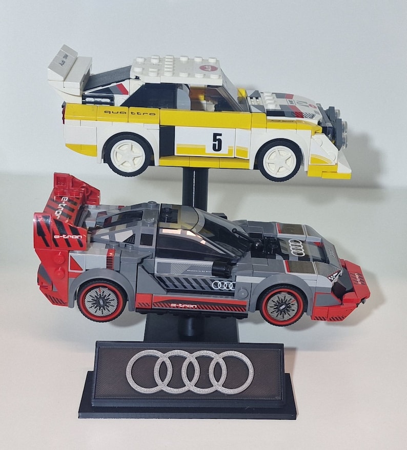 May include: Two Lego model race cars, one yellow and white with the number 5, and one gray and red, are displayed on a black stand. The stand features the Audi logo. The cars are detailed with racing decals.