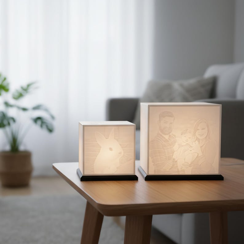 May include: Two illuminated cube lamps with black bases sit on a wooden table. One lamp displays a family portrait, the other a rabbit. The lamps are white and emit a soft glow, creating a warm ambiance.