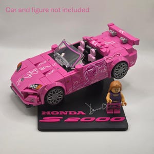 May include: A pink brick-built convertible sports car with white graphic decals, a clear windshield, and silver wheels. A small plastic figure with long brown hair stands on a black display base labeled HONDA S2000 in pink text.