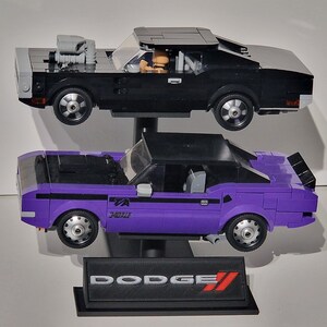 May include: Two Lego model cars, a black muscle car with a gray roof and a purple car with a black roof, are displayed. The purple car has the text "TA 340 Six" on the side. A black base with the Dodge logo is at the bottom.