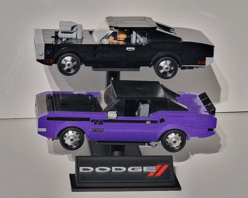 May include: Two Lego model cars, a black muscle car with a gray roof and a purple car with a black roof, are displayed. The purple car has the text "TA 340 Six" on the side. A black base with the Dodge logo is at the bottom.