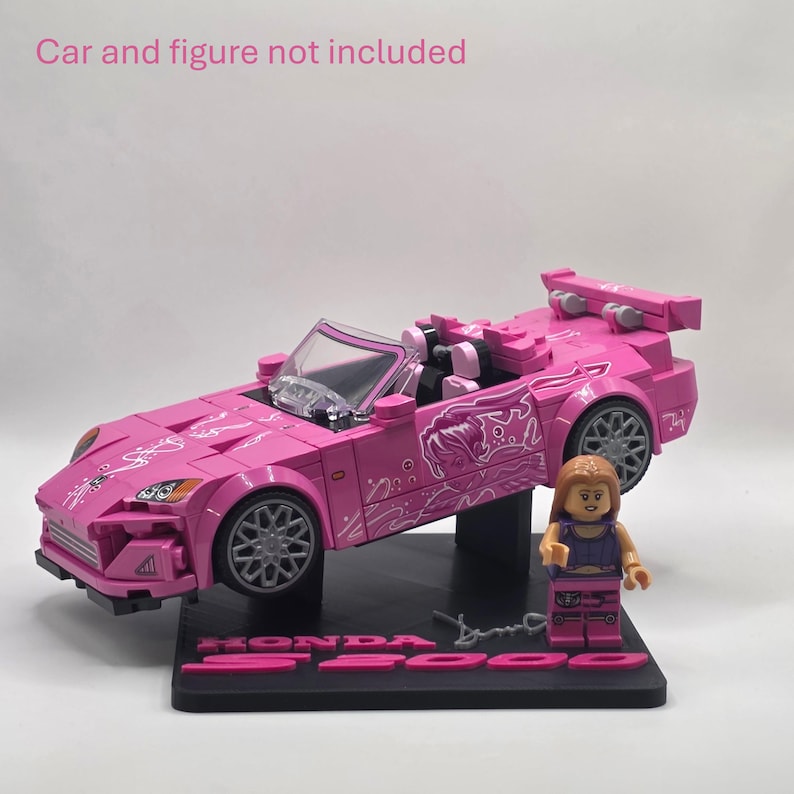 May include: A pink plastic building block model of a Honda S2000 convertible car with white graphic decals, a clear windshield, and grey wheels, mounted on a black display base with the text "HONDA S2000" and a small plastic figure.