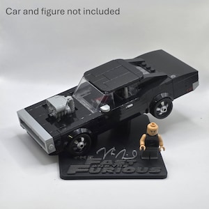 May include: A black brick-built muscle car model with a grey front bumper and exposed engine block, displayed on a black stand featuring the text "The Fast and the Furious" and a signature. A small plastic figure stands next to the car on the base.