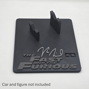 May include: A black rectangular display stand featuring two upright supports and the embossed text THE FAST AND THE FURIOUS with a stylized signature above it. The base includes a small two-dot detail and has a matte finish with visible print lines.