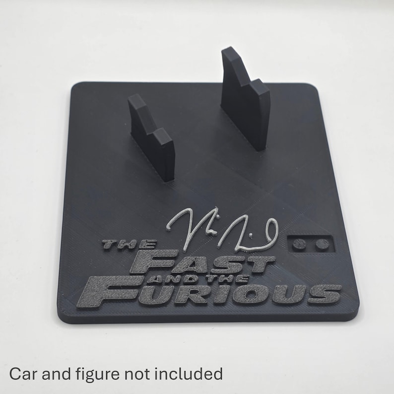 May include: A black rectangular display stand featuring two upright supports and the embossed text THE FAST AND THE FURIOUS with a stylized signature above it. The base includes a small two-dot detail and has a matte finish with visible print lines.