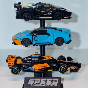 Puede incluir: Un expositor LEGO Speed Champions negro, azul y naranja con tres coches de carreras. El coche de arriba es negro con detalles dorados, el coche del medio es azul con detalles naranjas y el coche de abajo es naranja con detalles negros. El expositor tiene una base negra con las palabras "Speed Champions" impresas en &eacute;l.