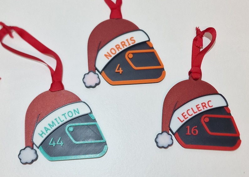 Op de afbeelding: Drie kerstversieringen in de vorm van racehelmen, elk met een kerstmuts. De ornamenten zijn rood, zwart en wit, met de namen "NORRIS", "HAMILTON" en "LECLERC" en hun racenummers.
