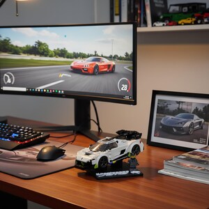May include: A white and black Lego sports car model with a black display stand, a Lego figure, and a computer monitor displaying a racing game. A framed car photo and a stack of magazines are also on the desk.