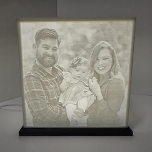 May include: A lighted photo cube displaying a family portrait. The cube features a black base and a glowing, etched image of a man, woman, and baby. The family is smiling. The cube is a personalized home decor item.