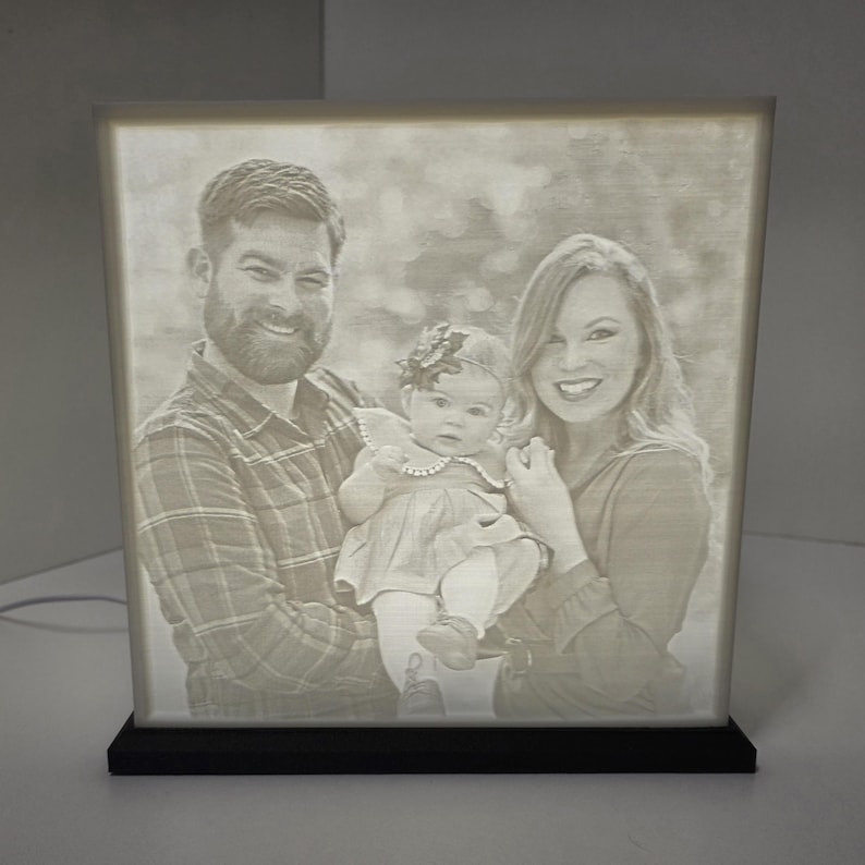 May include: A lighted photo cube displaying a family portrait. The cube features a black base and a glowing, etched image of a man, woman, and baby. The family is smiling. The cube is a personalized home decor item.