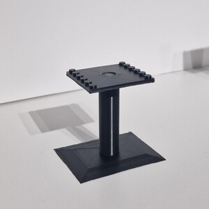 May include: A black plastic stand with a square top and a round hole in the center. The stand has a square base and a cylindrical column.