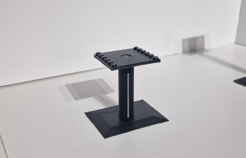 May include: A black plastic stand with a square top and a round hole in the center. The stand has a square base and a cylindrical column.