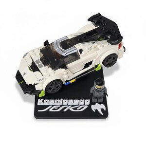 May include: A white and black toy sports car with a black spoiler and wheels. The car sits on a black base with the text "Koenigsegg Jesko" in white. A small figure in a gray suit and helmet stands next to the car.