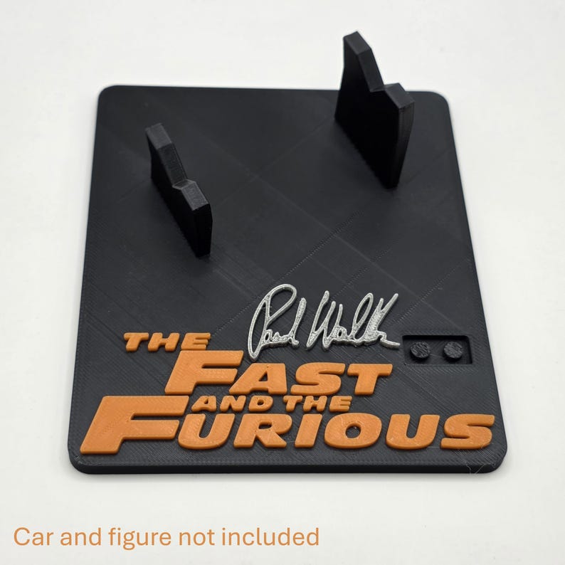 Pu&ograve; includere: Un espositore rettangolare nero con due supporti verticali, il testo arancione in rilievo "THE FAST AND THE FURIOUS", una firma color argento di Paul Walker e due piccoli perni circolari sulla superficie per scopi di montaggio.
