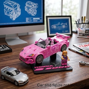 May include: A pink convertible toy car model with white graphic decals, a clear windshield, and silver wheels, mounted on a black display base labeled Honda S2000. A small plastic figure with long brown hair stands next to the car on the base.