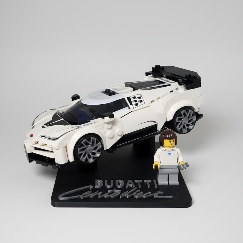 May include: A white and black Lego Bugatti Centodieci model car with a black base that reads "Bugatti Centodieci". A Lego figure stands next to the car. The car has black wheels and a black spoiler.