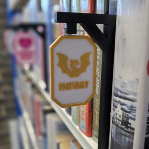 Op de afbeelding: Een wit en geel bord met het woord "FANTASY" en een draakafbeelding, bevestigd aan een zwarte metalen beugel. Het bord wordt weergegeven op een boekenplank tussen verschillende boeken.