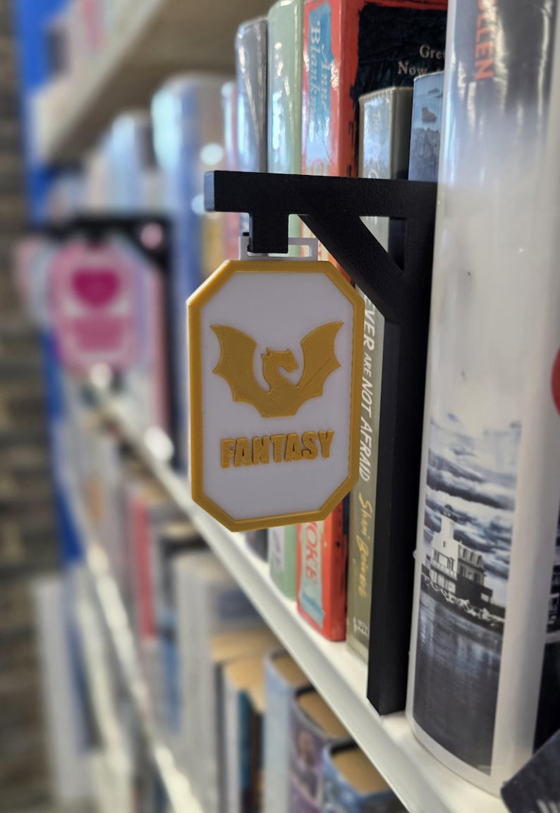 Op de afbeelding: Een wit en geel bord met het woord "FANTASY" en een draakafbeelding, bevestigd aan een zwarte metalen beugel. Het bord wordt weergegeven op een boekenplank tussen verschillende boeken.