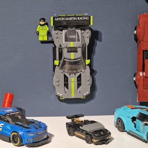 Puede incluir: Una colecci&oacute;n de cuatro coches de carreras Lego, incluyendo un coche Aston Martin Racing gris y verde, un coche negro y dorado, un coche azul y negro y un coche rojo. El coche Aston Martin Racing tiene una figura Lego de un piloto de carreras de pie junto a &eacute;l.