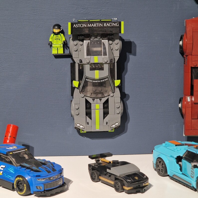 Puede incluir: Una colecci&oacute;n de cuatro coches de carreras Lego, incluyendo un coche Aston Martin Racing gris y verde, un coche negro y dorado, un coche azul y negro y un coche rojo. El coche Aston Martin Racing tiene una figura Lego de un piloto de carreras de pie junto a &eacute;l.