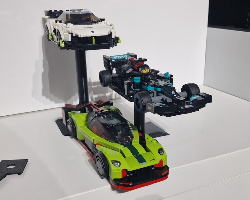 May include: Three toy race cars on display stands. A white and green car is on the top stand, a black and blue car with the word "PETRONAS" on the side is on the middle stand, and a green and black car is on the bottom stand.