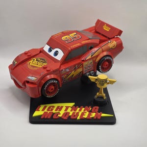 Puede incluir: Modelo de coche de carreras rojo Lightning McQueen con el n&uacute;mero 95 en el cap&oacute; y los laterales. El modelo est&aacute; sobre una base negra con las palabras "LIGHTNING MCQUEEN" en amarillo y rojo. Tambi&eacute;n hay un trofeo de la Copa Pist&oacute;n dorado.