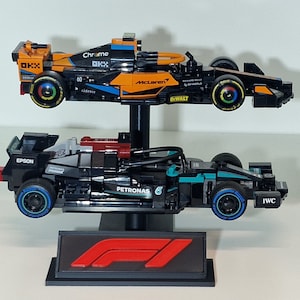 May include: Two Formula 1 race car models, one orange and black, the other black and teal, are displayed on a black stand. The orange car has "McLaren" and "Chrome" branding. The black car has "Petronas" and "IWC" branding. A red and black F1 logo is at the base.