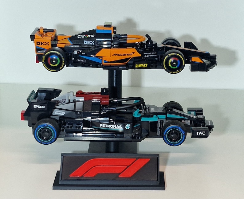 May include: Two Formula 1 race car models, one orange and black, the other black and teal, are displayed on a black stand. The orange car has "McLaren" and "Chrome" branding. The black car has "Petronas" and "IWC" branding. A red and black F1 logo is at the base.