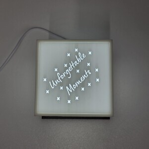 May include: Square, illuminated sign with the words "Unforgettable Moments" in white script, surrounded by small star shapes. The sign has a white border and a black base, with a power cord extending from the top.