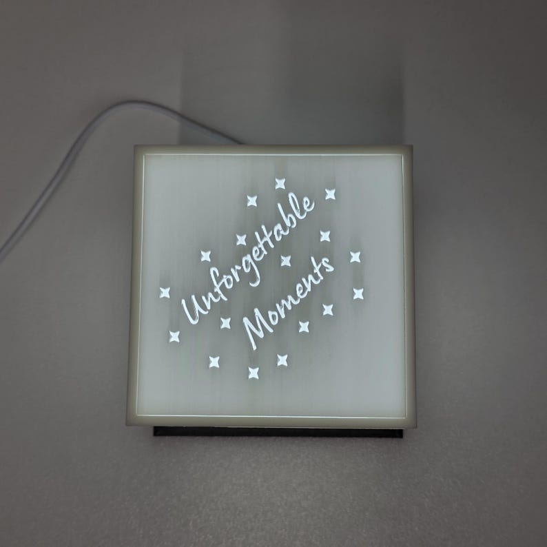 May include: Square, illuminated sign with the words "Unforgettable Moments" in white script, surrounded by small star shapes. The sign has a white border and a black base, with a power cord extending from the top.
