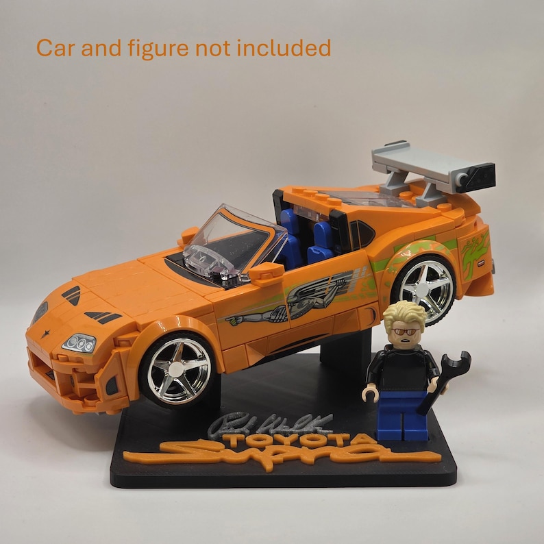 Pu&ograve; includere: Un modellino di auto sportiva Toyota Supra arancione realizzato con mattoncini, dotato di alettone posteriore, ruote argentate e grafiche laterali verdi. Una piccola figura in plastica con capelli biondi e maglia nera su una base nera con scritta.