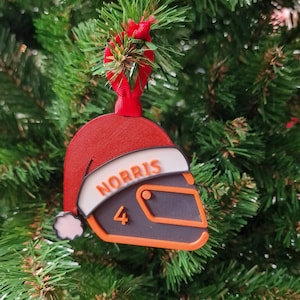 Puede incluir: Adorno navideño con forma de casco de carreras, con diseño de gorro de Papá Noel. El casco es gris oscuro con detalles en naranja, con el nombre "NORRIS" y el número "4". Cuelga de una cinta roja.