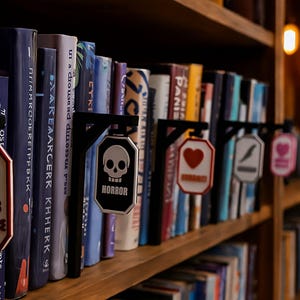 Op de afbeelding: Een boekenplank met diverse boeken en decoratieve boekenleggers. De markers hebben labels zoals "Sports Romance" met een voetbalafbeelding, "Horror" met een schedel en een hartsymbool. De boeken zijn in verschillende kleuren.
