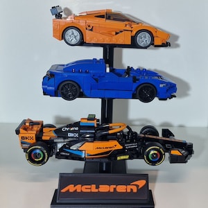 Puede incluir: Un expositor de tres niveles con tres coches de carreras McLaren. El coche de arriba es naranja, el del medio es azul y el de abajo es naranja y negro con las palabras "Chrome", "McLaren" y "Dewalt" en &eacute;l. El expositor tiene una base negra con la palabra "McLaren" en ella.