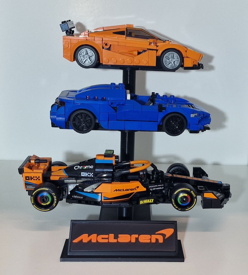 Puede incluir: Un expositor de tres niveles con tres coches de carreras McLaren. El coche de arriba es naranja, el del medio es azul y el de abajo es naranja y negro con las palabras "Chrome", "McLaren" y "Dewalt" en &eacute;l. El expositor tiene una base negra con la palabra "McLaren" en ella.