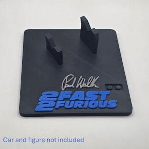 May include: A black rectangular display stand featuring two upright supports, a silver-colored signature of Paul Walker, and the blue text 2 FAST 2 FURIOUS. The base includes a small rectangular cutout with two circular pegs. Car and figure not included.